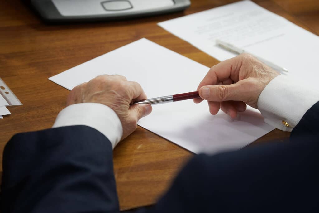 Elderly Man In A Strict Business Suit, Sitting At A Table With A